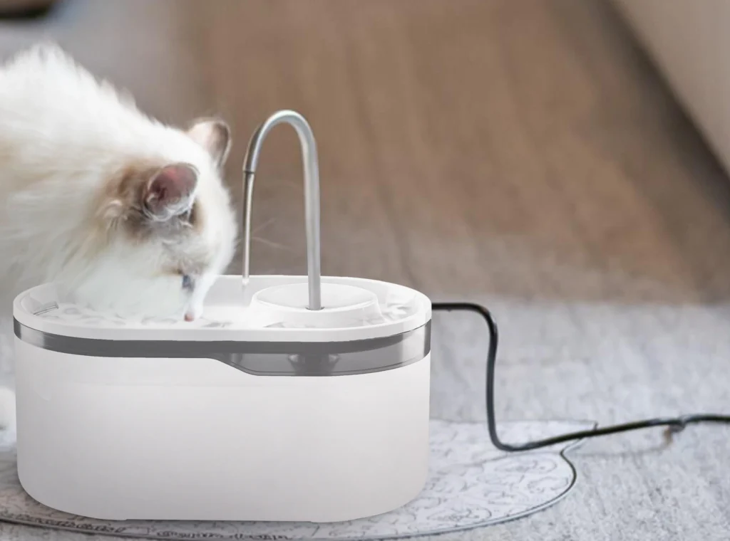 A fluffy Ragdoll cat drinking from a white plastic automatic cat water fountain with a curved metal spout, showing a popular customer OEM model.