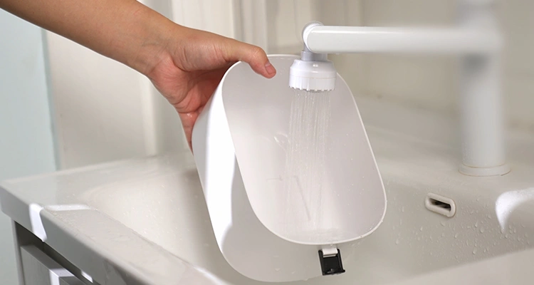A hand rinsing the white plastic water reservoir of a pet fountain under running water in a sink, showing proper hygiene to avoid a dirty cat water fountain.